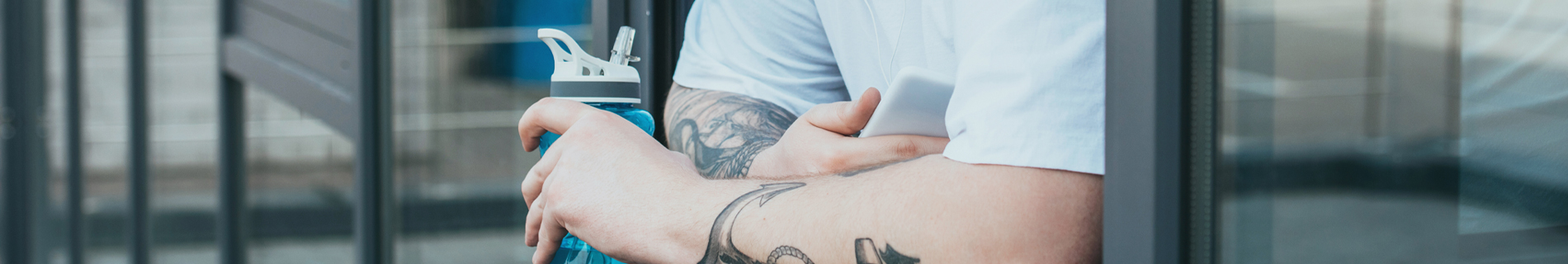 Man with tattooed arms leaning against the window holding his cell phone and a bottle of water.